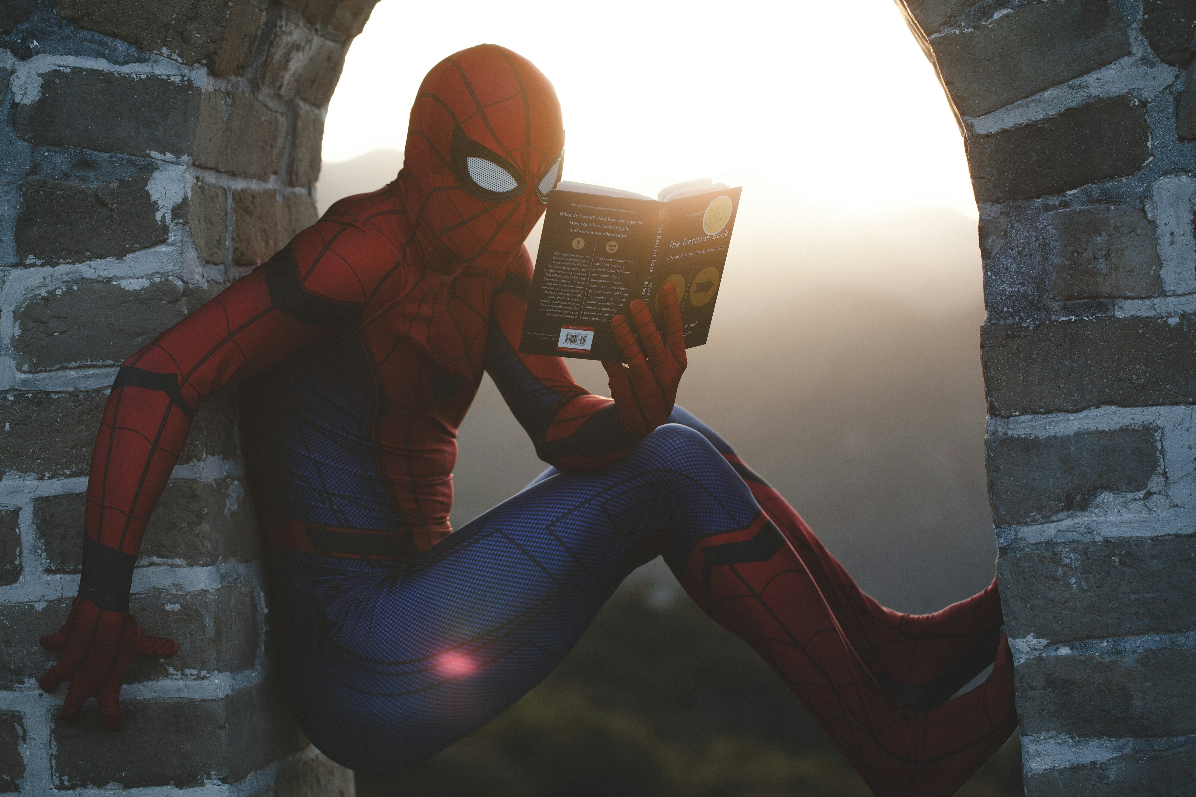 A person wearing a Spiderman costume sits in an open window reading a book.