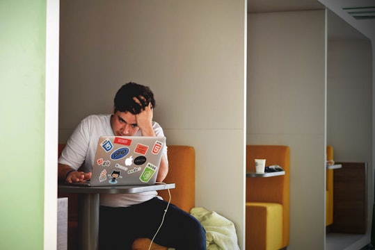 A man in a white t-shirt looks works at a laptop, holding his head in frustration.