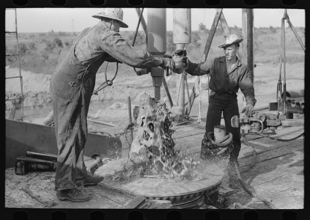 black and white photo of two oil workers working on a pipe with oil spilling from it