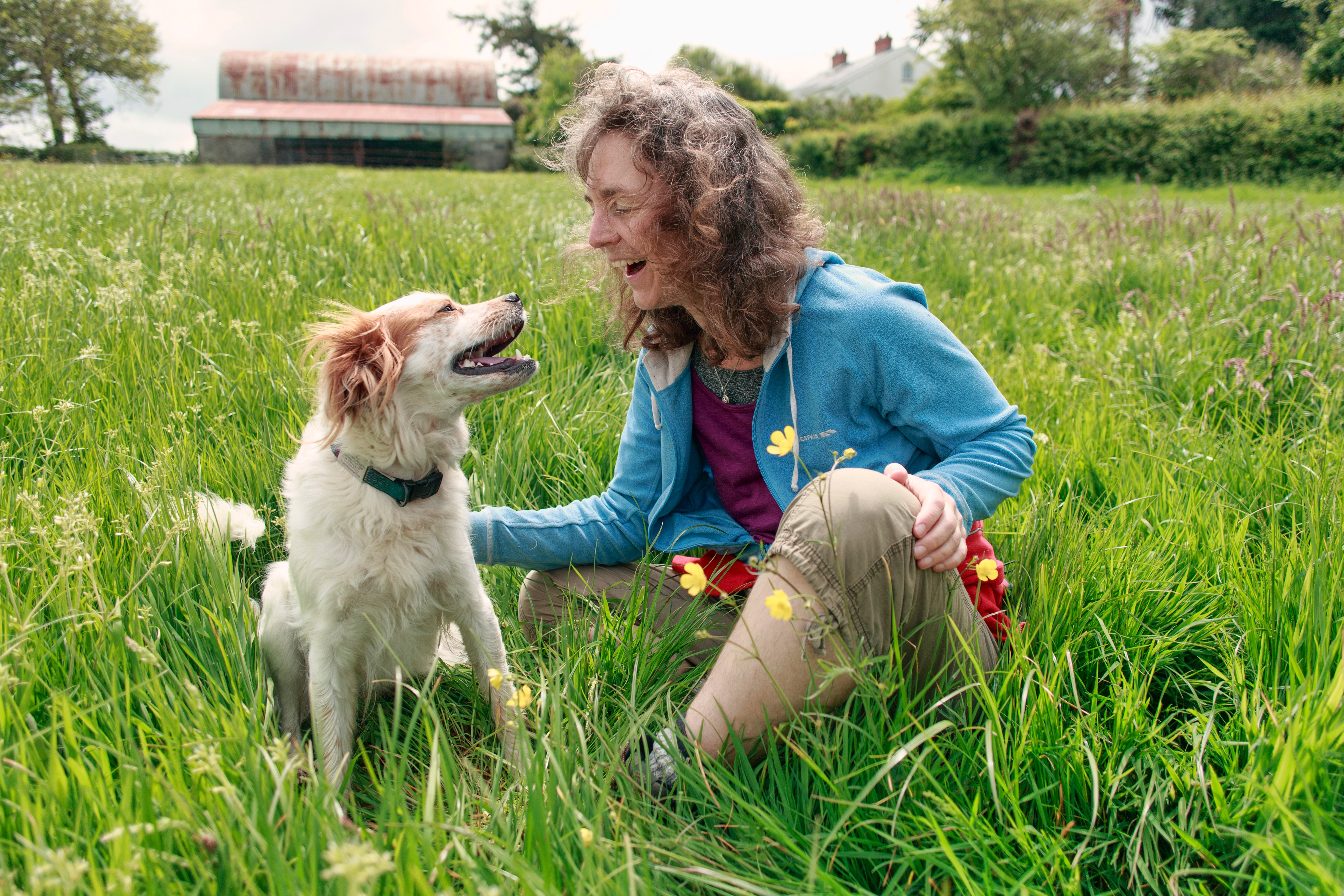 a woman sitting in the grass smiling at her dog, who is smiling back
