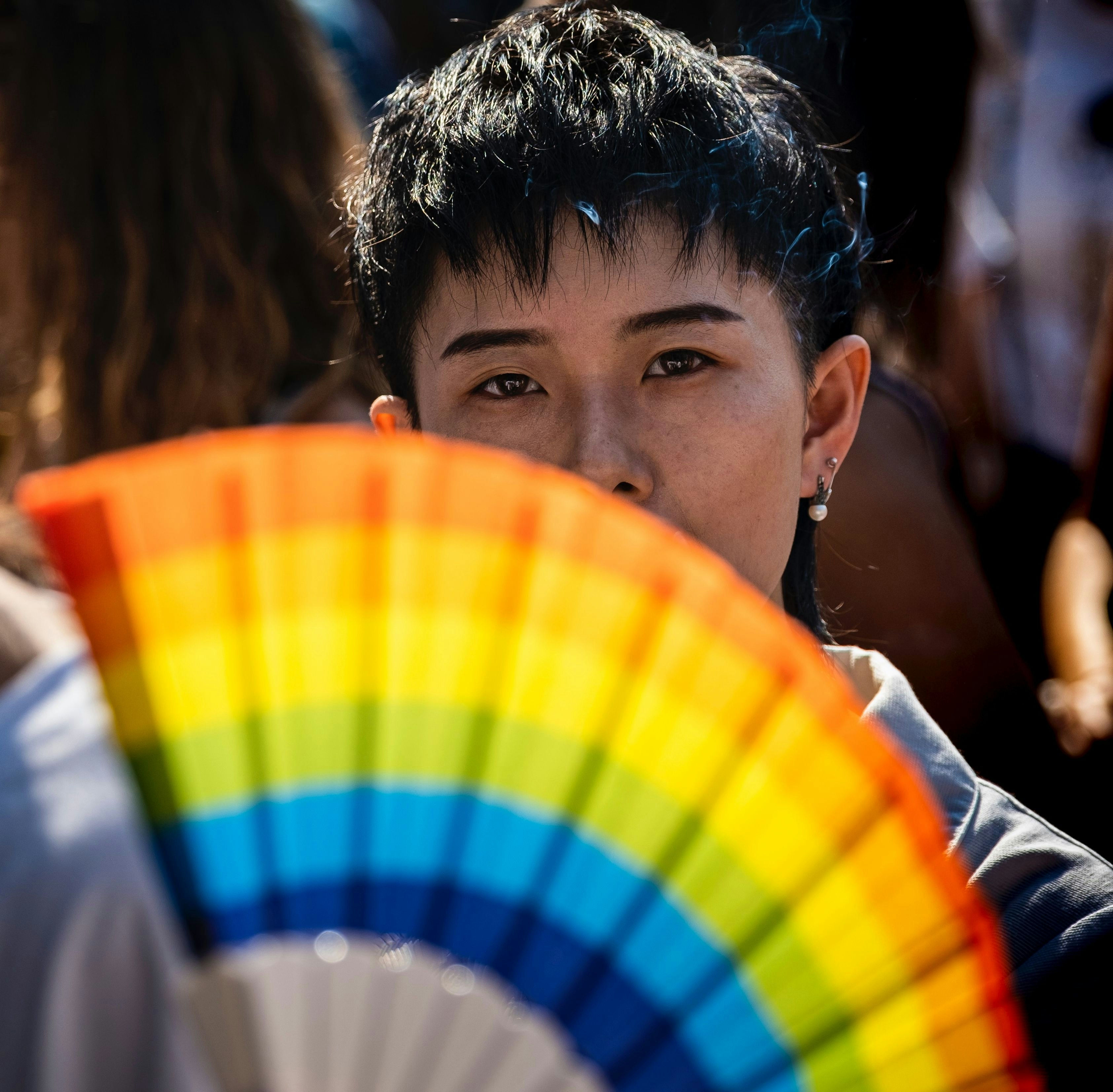A person looks out from behind a rainbow-colored fan.