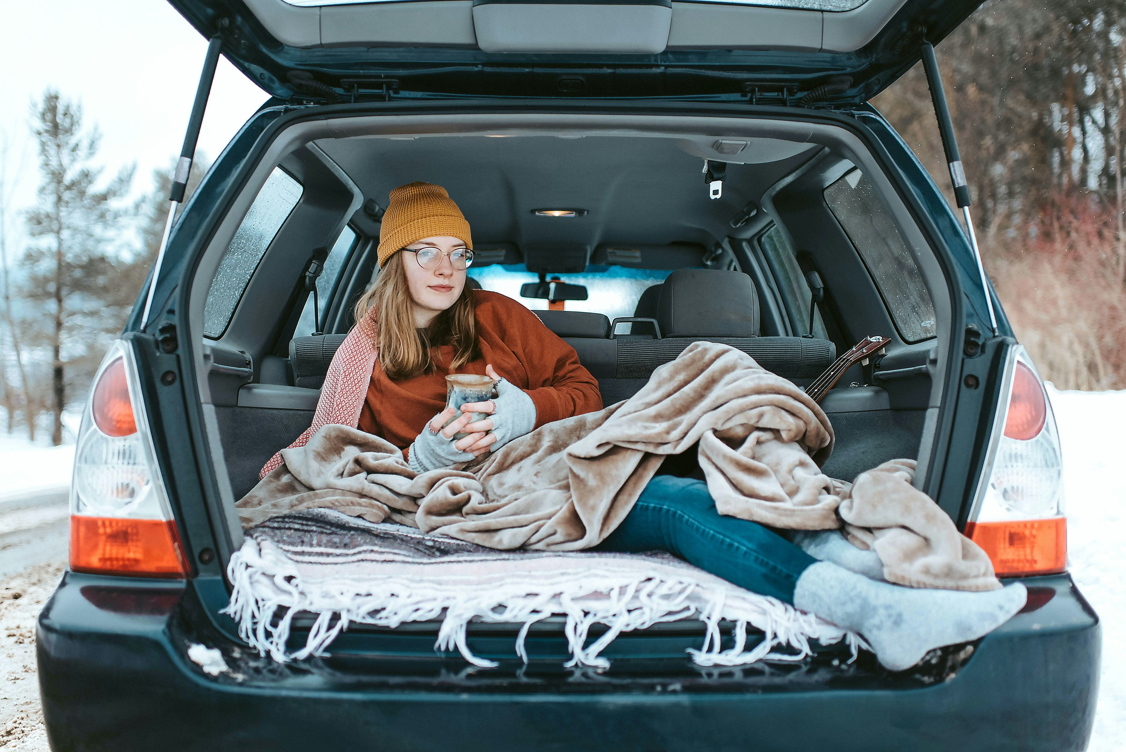 A woman sitting the trunk of a car drinking coffee, under a blanket