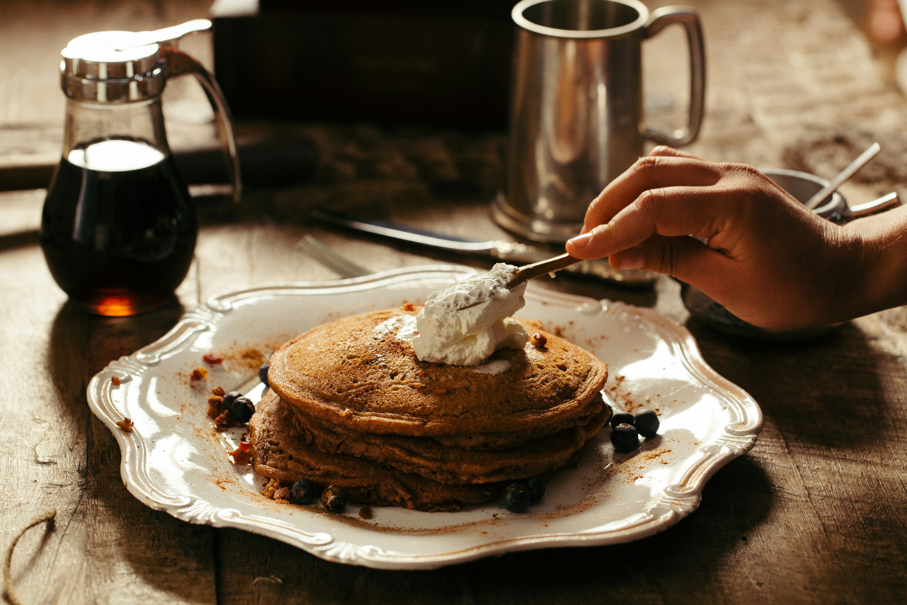 A person spreading butter on a stack of pancakes, with syrup and coffee next to the plate.