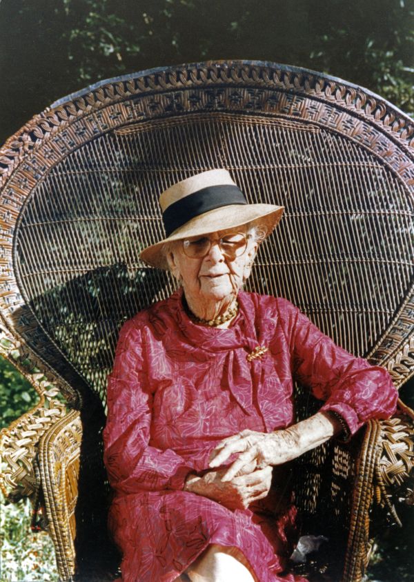 Marjory Stoneman Douglas, wearing a red dress and a brimmed sun hat, sitting in a high backed wicker chair.