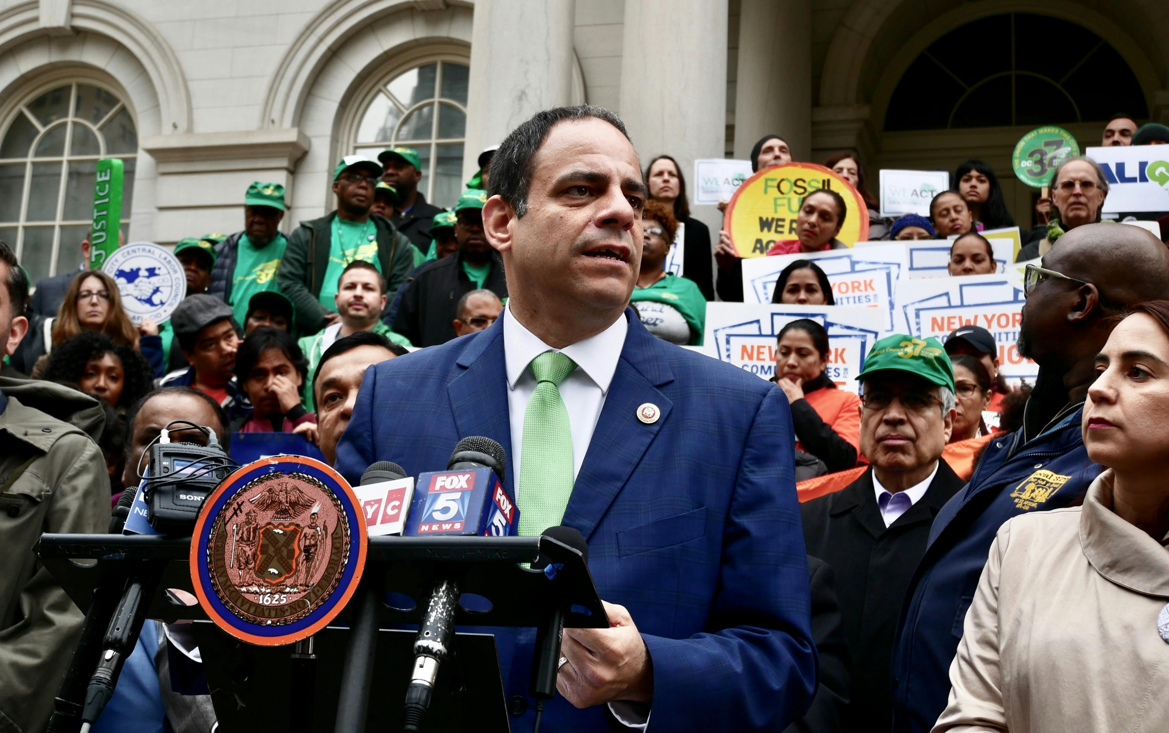Costa Constantinides speaks at a podium in front of a crowd of people at a rally for climate legislation. They are standing in front of New York City Hall.