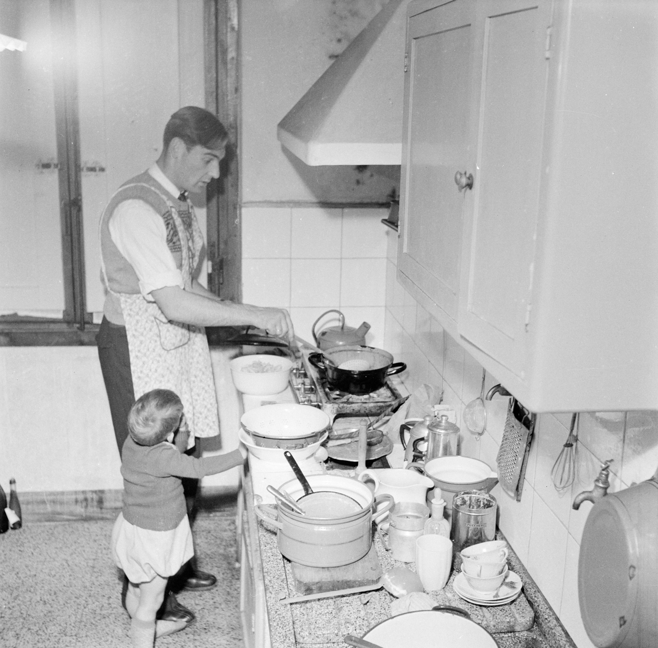 A black and white photo of a man cooking in a cluttered kitchen, with a small child tugging at his apron.
