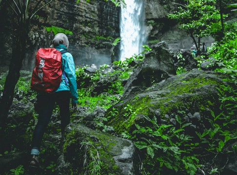 a woman wearing a hijab with a red backpack hiking to a waterfall