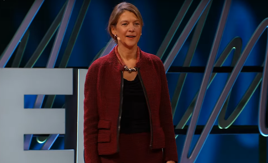 heidi larson, a woman wearing a red suit, standing on stage to give a talk