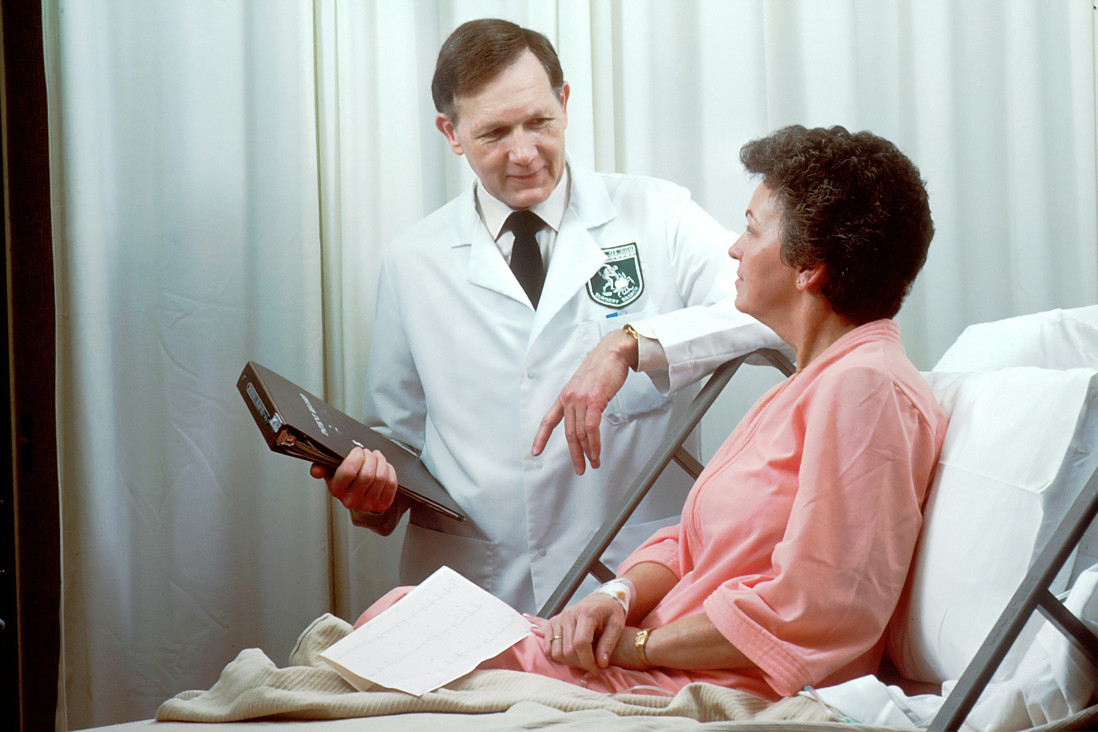 A white doctor standing over and speaking with a white patient laying in a hospital bed