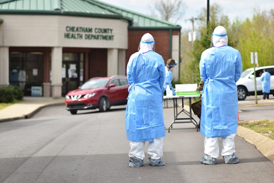 Two soldiers wearing personal protective equipment (PPE) stand waiting for a car in a drive-through coronavirus testing location. 