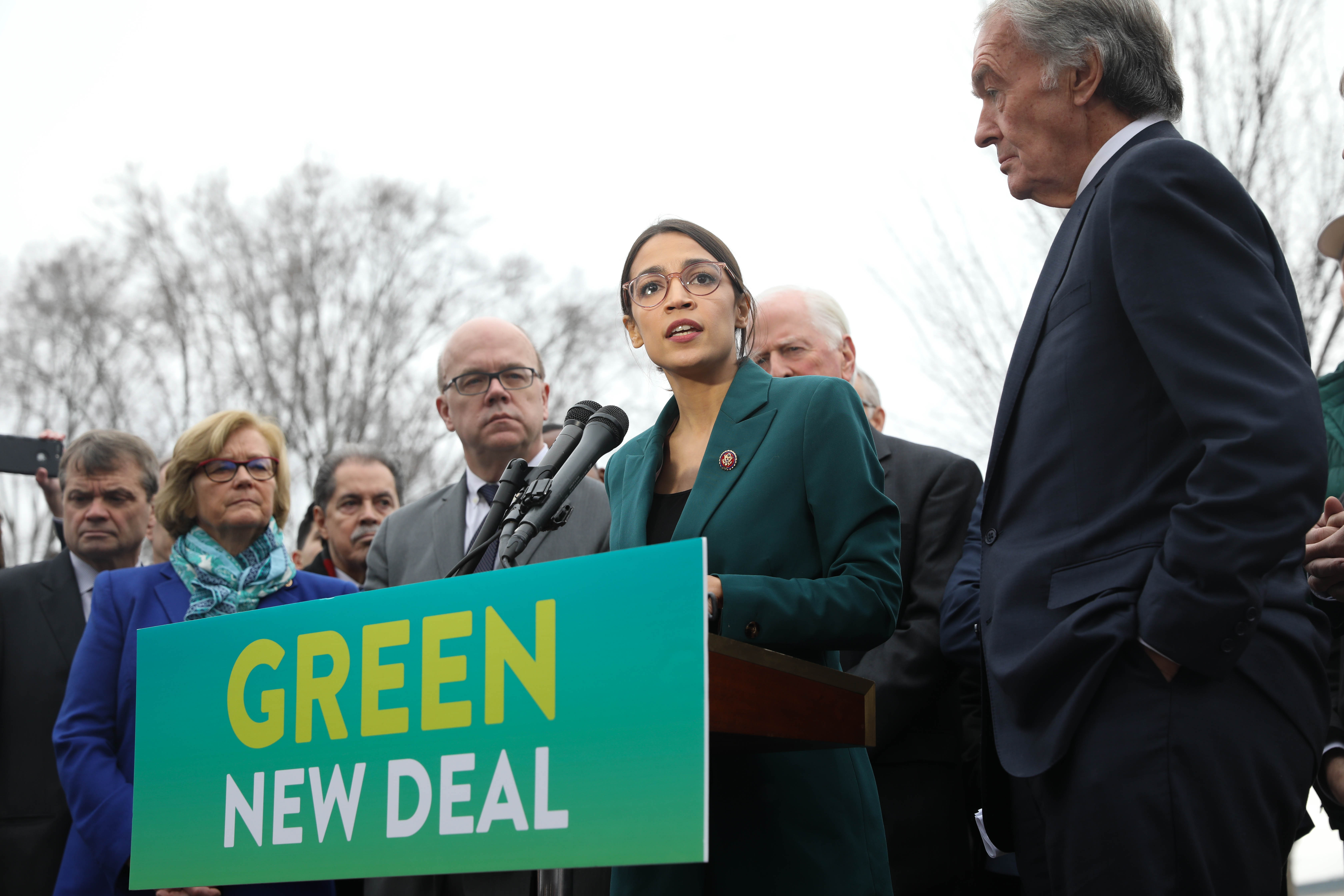 Senate and House Democrats stand at a podium while Representative Alexandria Ocasio-Cortez speaks into a microphone about the Green New Deal and climate change legislation.