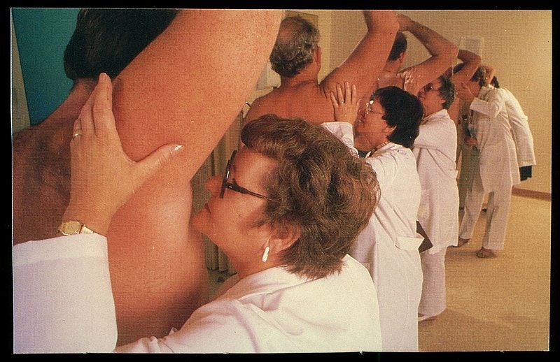 a line of women wearing lab coats smelling the armpits of a bunch of men