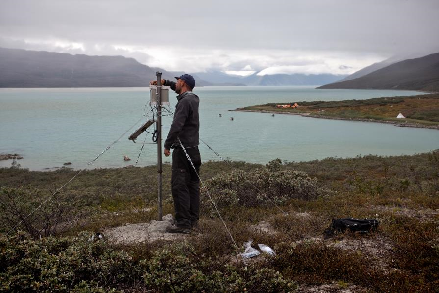 Next to a lake ringed by mountains, a scientist sets up a surveying probe.
