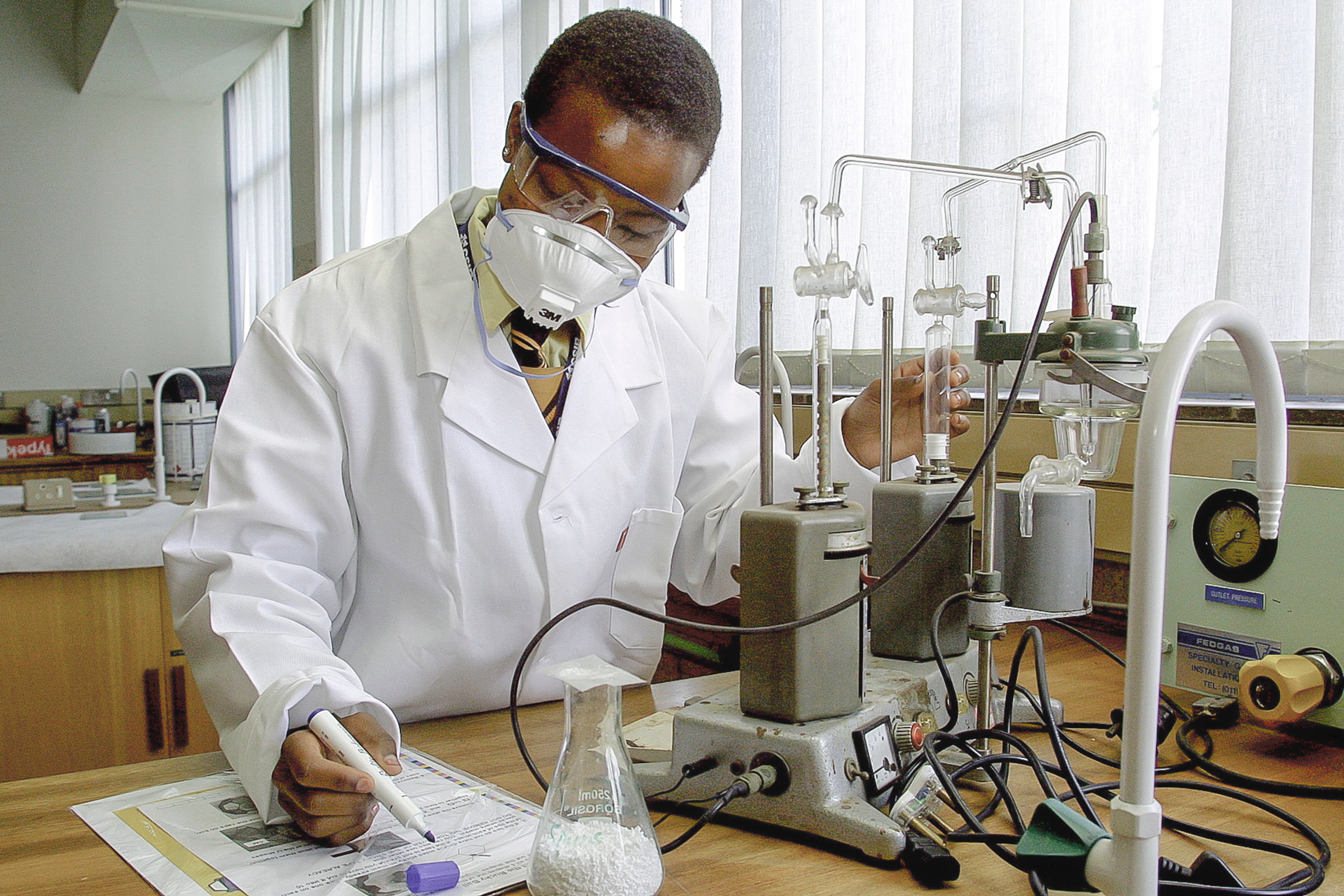 A scientist, wearing a lab coat and a face mask, takes notes on an experiment.