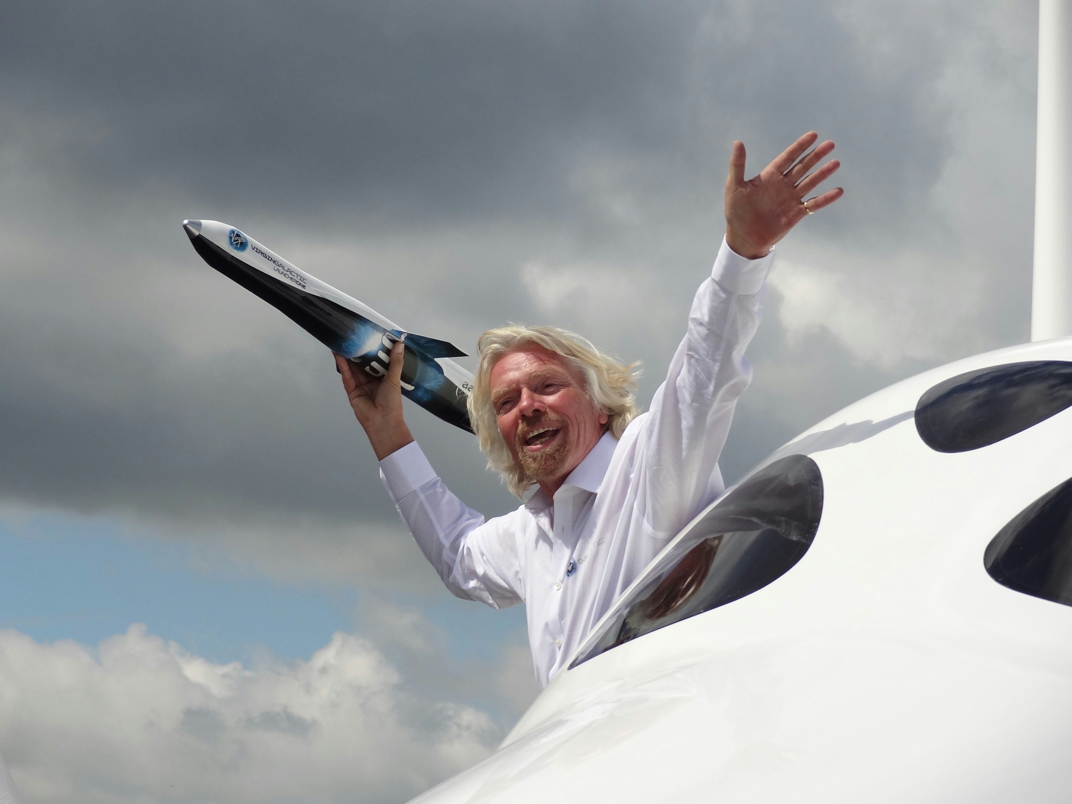 richard branson hanging out of an airplane holding a small replica spaceship