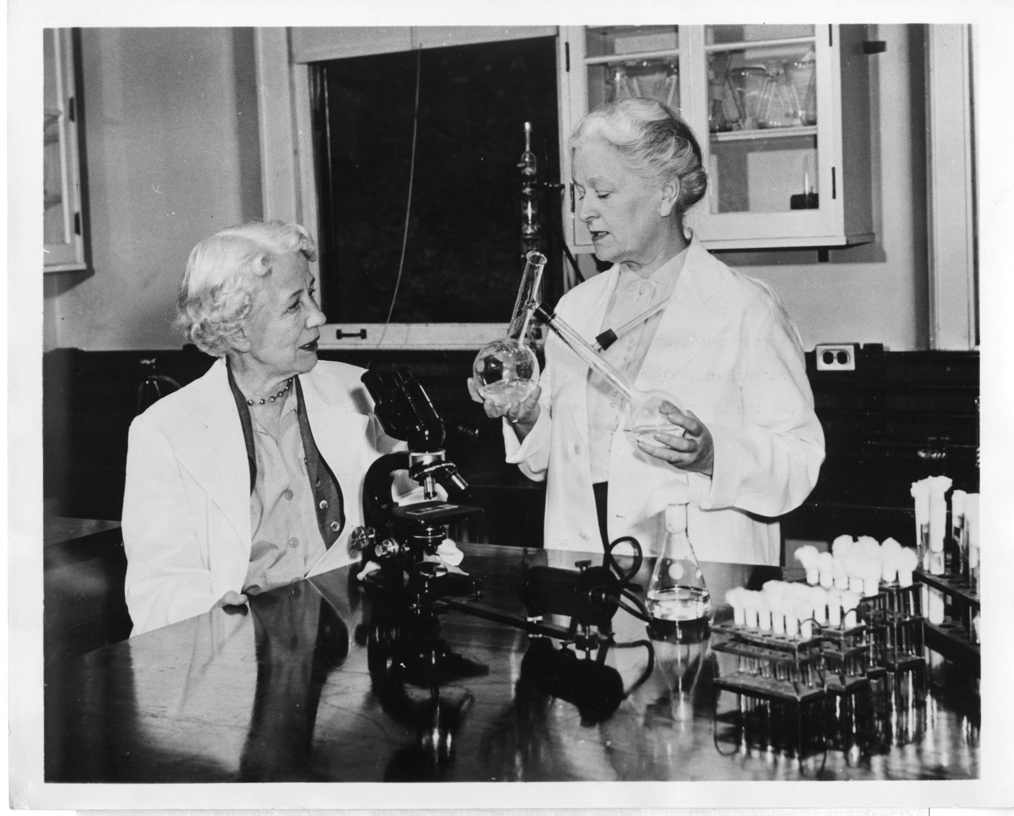 Microbiologist Elizabeth Lee Hazen and chemist Rachel Brown, standing at a table with a microscope and culture tubes.