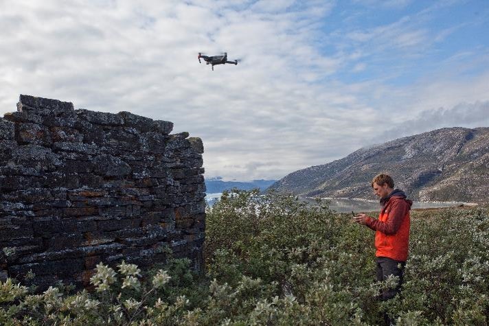 A scientist pilots a drone over the ruins of a stone structure in a grassy field.