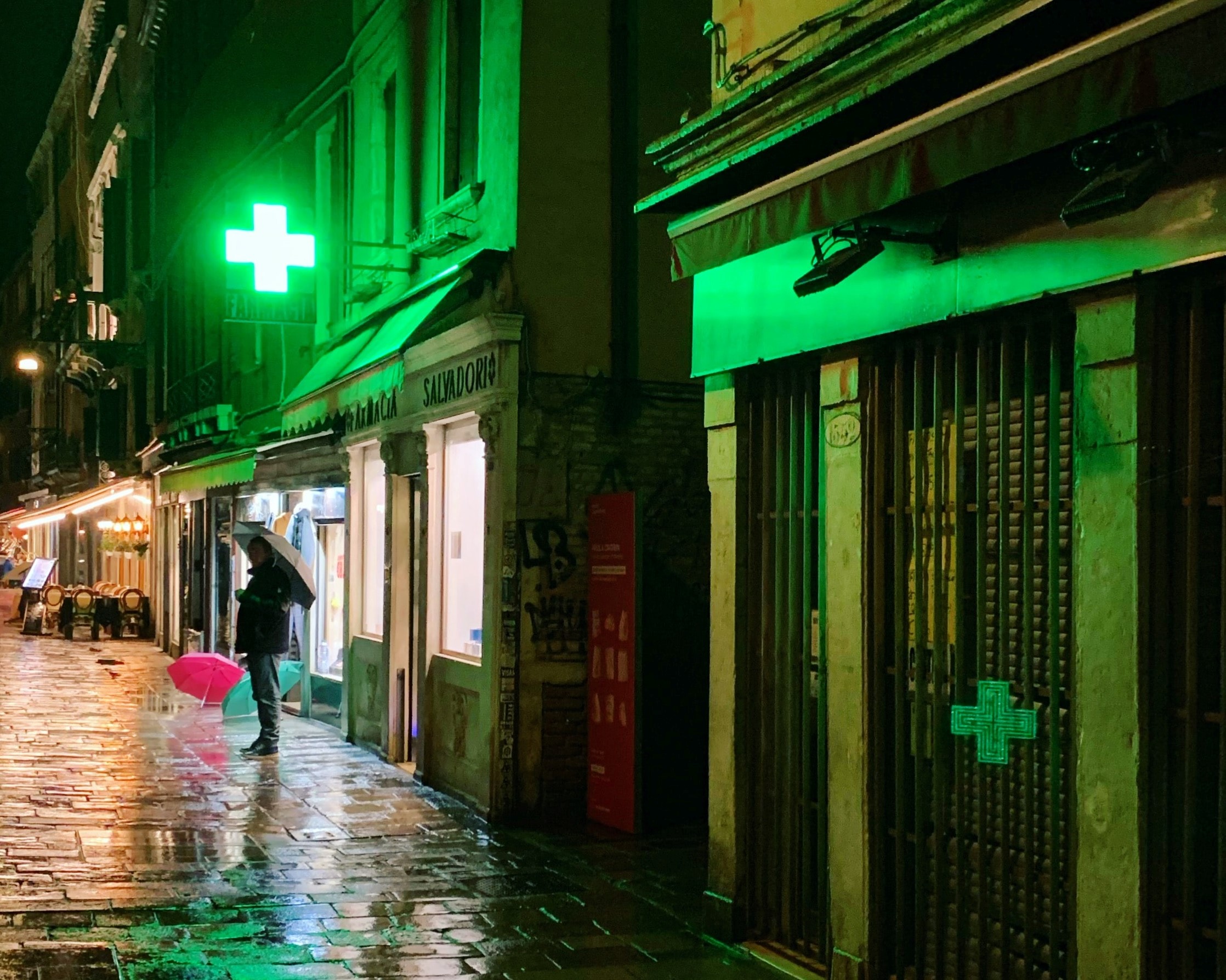 A pharmacy with a neon green cross sign on a rainy street at night