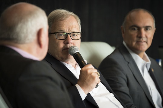 man seated next to two other men, wearing glasses and holding a microphone 