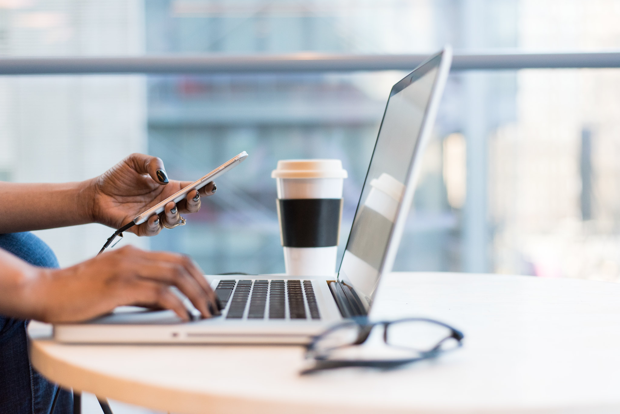 person holding a phone and working at a laptop with coffee and glasses