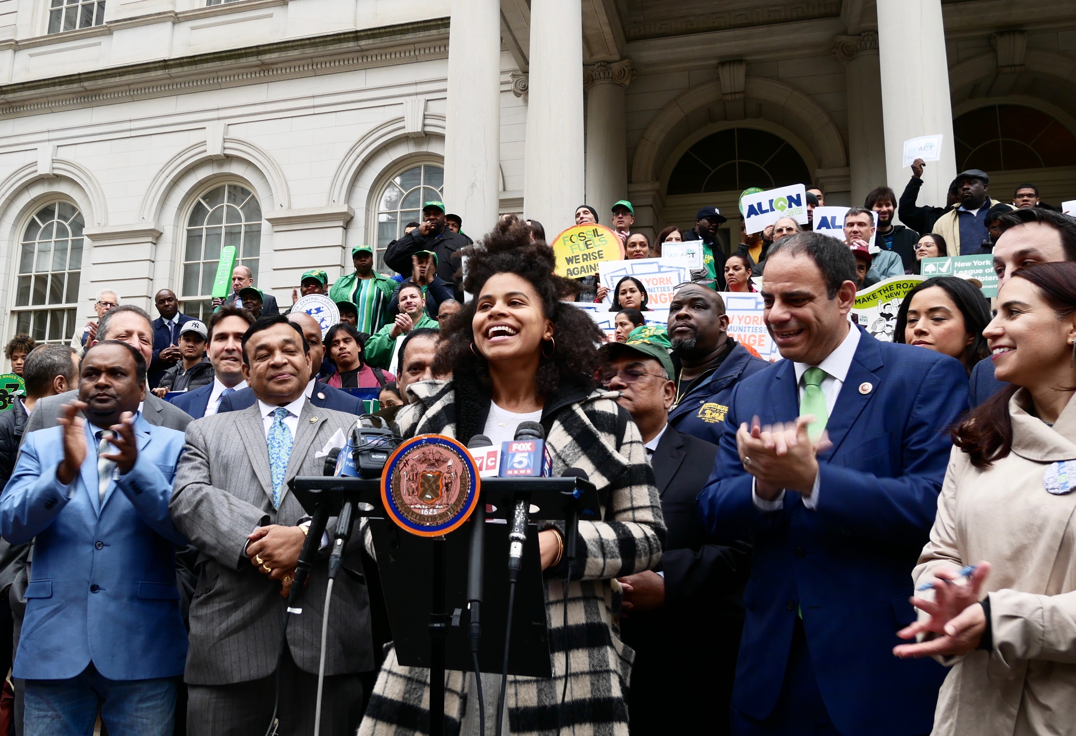 Zazie Beetz speaks in front of a podium and a crowd of people in front of New York City Hall in support of climate change legislation.