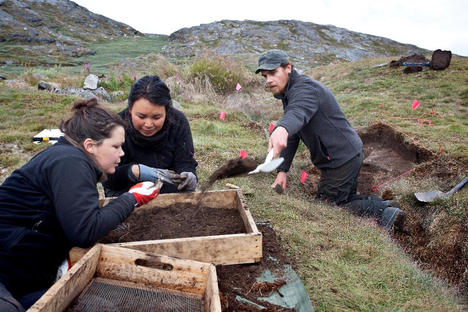 Three scientists digging at an archaeological site in Greenland. Two are inspecting a find while a third digs.