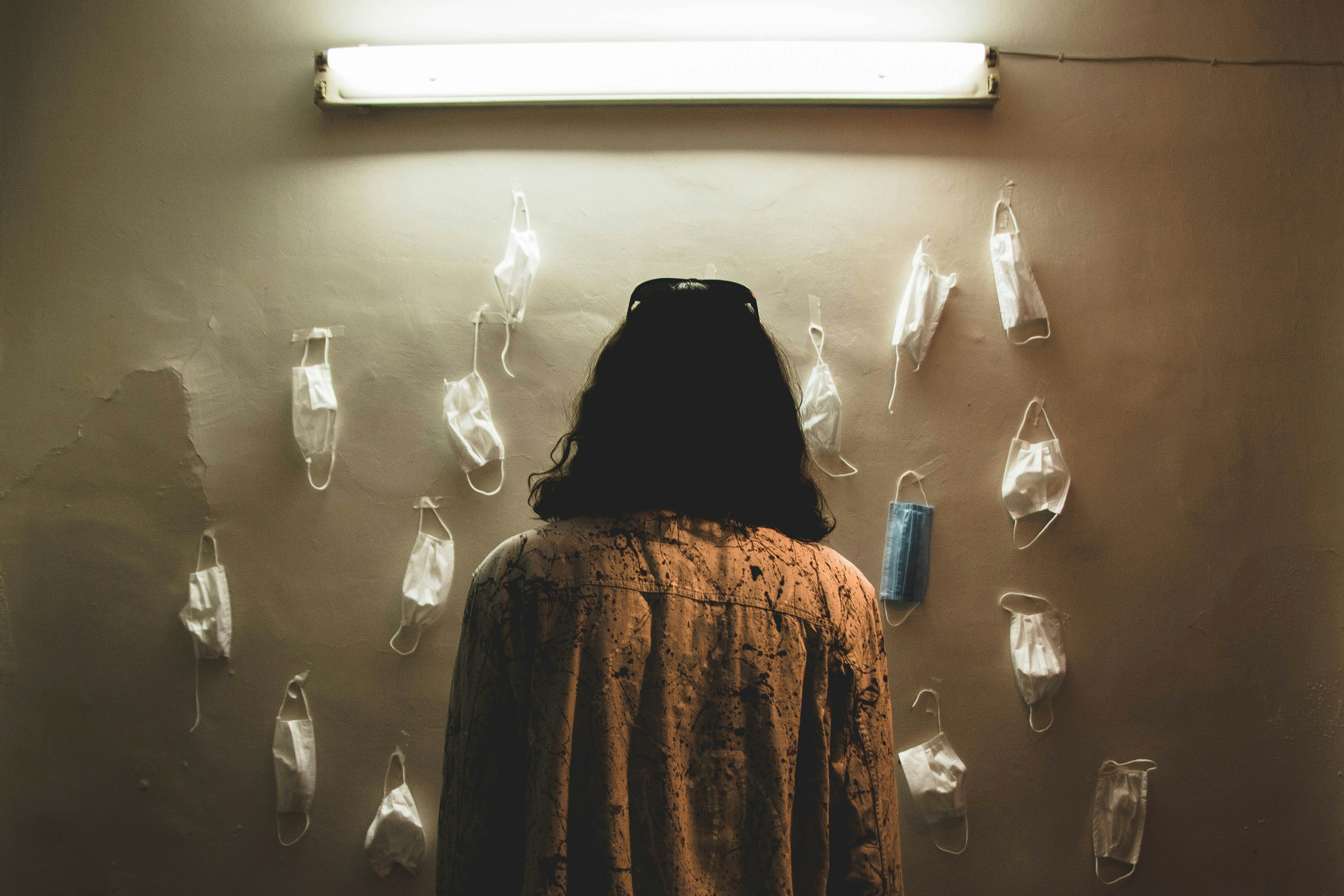 a woman staring at masks hanging on a wall. almost all masks are white, but one is blue