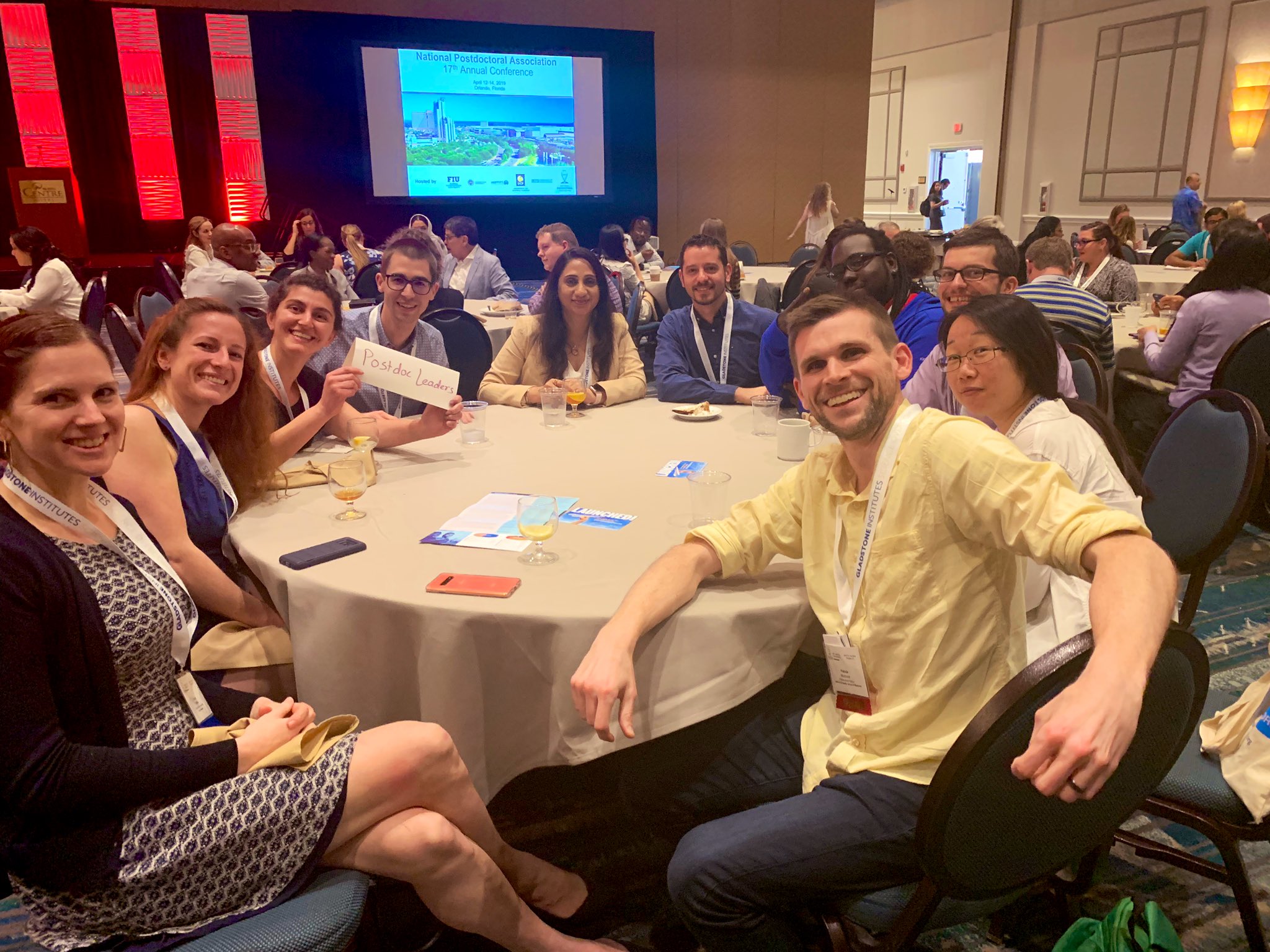 A group of people posing for a photo seated at a circular table. One is holding a paper sign that reads "Postdoc Leaders."