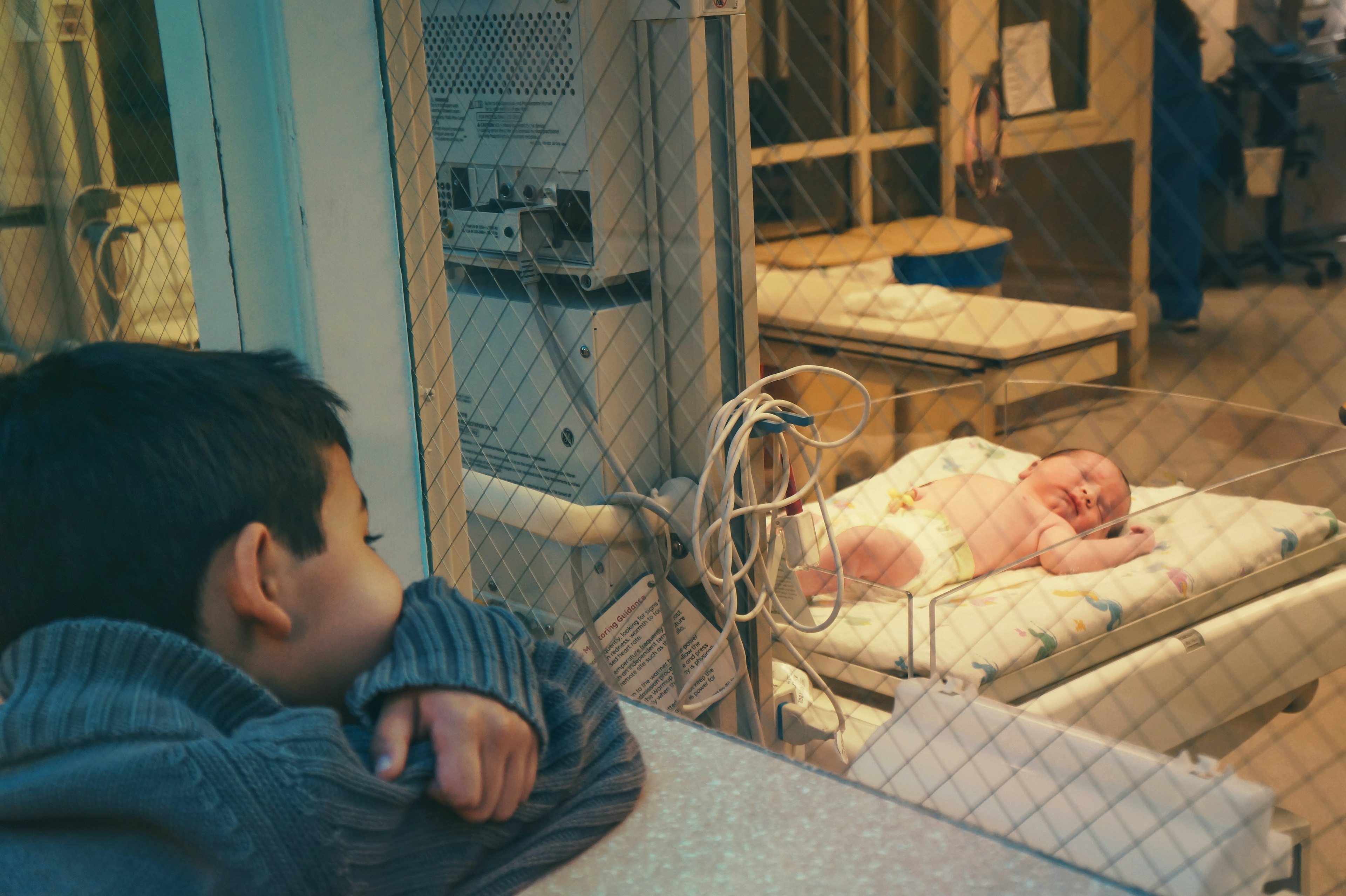 A child looks through a glass window at a newborn in a bassinet